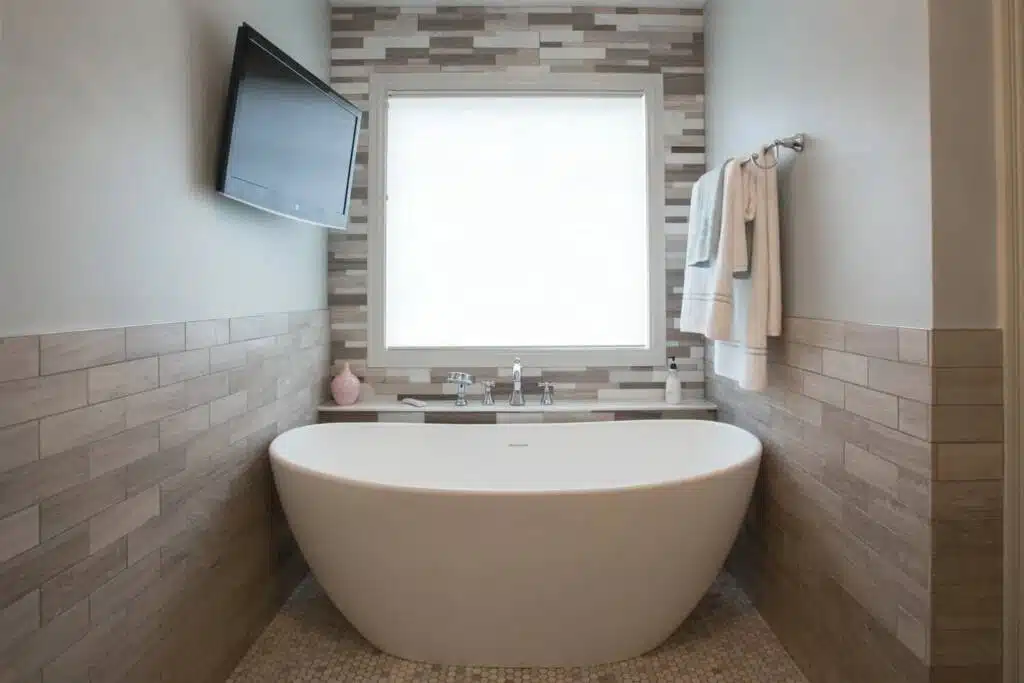 Tiled backdrop and TV mounted above a rounded oval bathtub in a compact bathroom remodeling project.